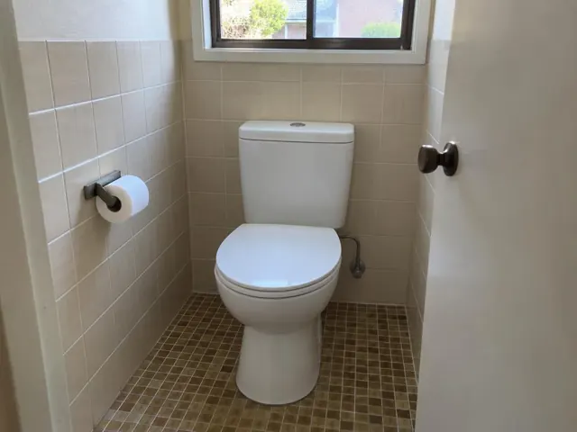 Clean white toilet in a tiled small bathroom with a window and open door in Brookvale